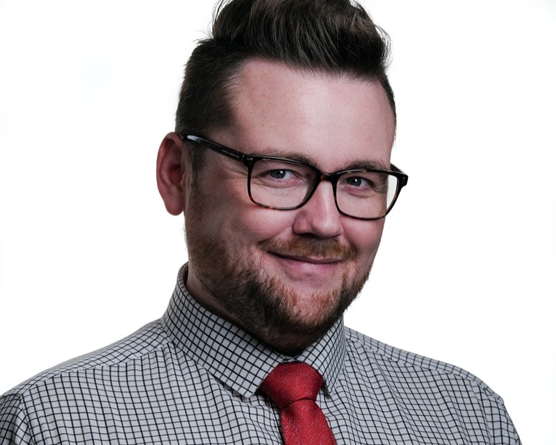 Headshot of a man with a short beard and glasses with a rectangular frame. He has a checked shirt with a red tie.