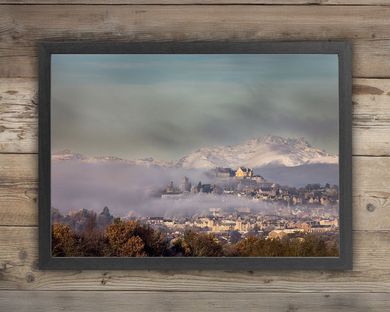 In a frame on a wooden background, we see a photo of clouds rolling over a town at the bottom of a hill. In the foreground, we look over trees, and in the background is a snowy mountain.